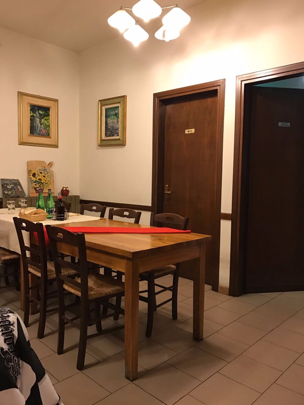 La sala interna della trattoria: tavoli in legno con tovaglia bianca e runner rosso, sedie rustiche impagliate